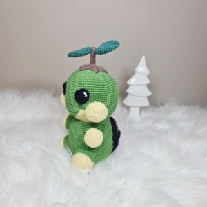 May include: A handmade crochet plush toy in the shape of a green turtle-like creature with a sprout on its head. The toy has black eyes, cream-colored accents, and a brown shell. It is sitting on a white, fluffy surface.