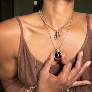 May include: A person wearing a brown tank top with two necklaces. The top necklace is a delicate chain with a small scorpion pendant. The bottom necklace is a thicker chain with a round pendant featuring a black and white yin yang symbol.