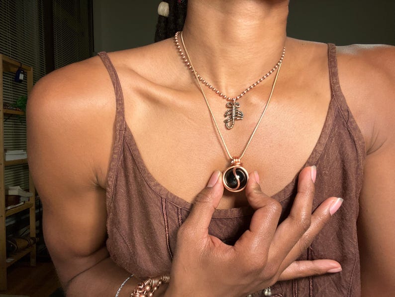 May include: A person wearing a brown tank top with two necklaces. The top necklace is a delicate chain with a small scorpion pendant. The bottom necklace is a thicker chain with a round pendant featuring a black and white yin yang symbol.