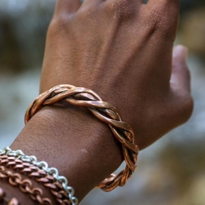 May include: A copper-coloured braided cuff bracelet. The bracelet is worn on a wrist, with other bracelets visible below. The braided design is the focal point, showcasing the warm tones of the metal. The image is well-lit, highlighting the texture and craftsmanship.