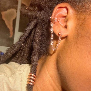 May include: A copper-colored ear cuff connected to a small hoop earring by a delicate chain. The ear cuff features a coiled wire design, while the chain consists of small, interlocking links. A matching copper-colored coil adorns a lock of hair.