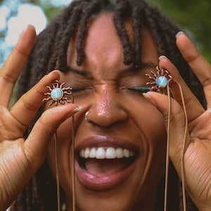 May include: A person with dreadlocks is holding up two gold necklaces with sun pendants featuring blue and white opalescent stones. The necklaces are hanging down in front of the person's chest.