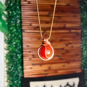 May include: A copper-colored wire-wrapped pendant necklace featuring a round, orange gemstone. The pendant hangs from a delicate gold-colored chain. The background includes a bamboo blind and greenery.
