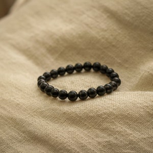 May include: A black beaded bracelet made of round, smooth stones. The bracelet is lying on a beige fabric surface.
