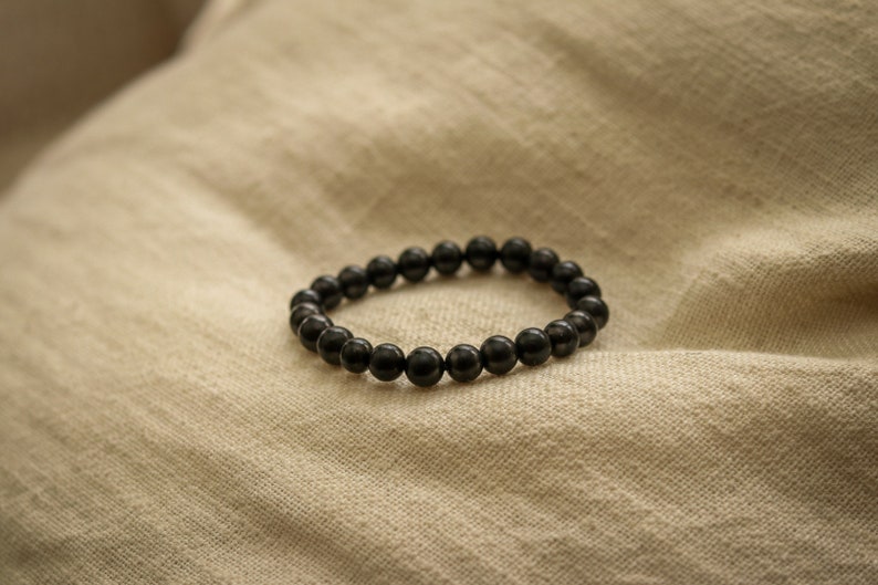 May include: A black beaded bracelet made of round, smooth stones. The bracelet is lying on a beige fabric surface.