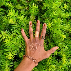 May include: A hand adorned with copper jewelry, including a ring, bracelet, and a spiral hand piece. The hand is set against a backdrop of vibrant green foliage, showcasing the jewelry's warm tones. The nails are painted with a metallic brown polish.