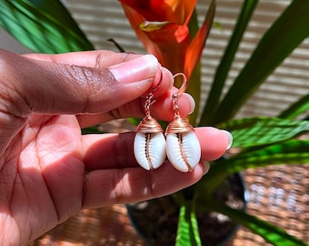 May include: A pair of gold-toned earrings featuring a cowrie shell wrapped in wire. The earrings are held in a hand against a blurred background of a green plant with orange flowers.