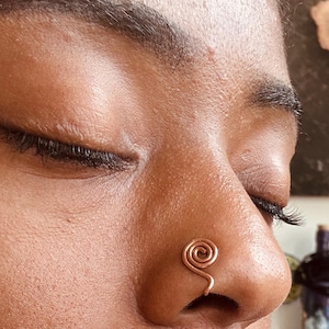 May include: A copper-colored nose cuff featuring a circular spiral design. The metal wire is coiled into a tight, round shape that sits securely on the side of the nostril, providing a decorative accent without the need for a piercing.