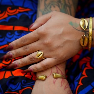 May include: Gold-coloured jewellery set featuring a ring and bracelets with a seashell design. The ring is on a finger, and the bracelets adorn the wrist. The background includes a blue and orange patterned fabric.