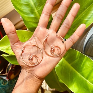 May include: Two sets of copper-toned hoop earrings, each featuring a larger hoop and a smaller hoop connected by a wrapped wire detail. The earrings have a metallic finish and are circular in shape, showcasing a simple design with wire accents.
