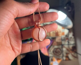 May include: A rose quartz pendant necklace with a copper wire cage. The pendant is a round, smooth stone with a light pink color. The necklace is made of a thin, gold-colored chain.