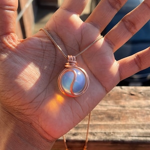 May include: A rose gold necklace with a wire-wrapped opal pendant. The opal is a light blue color with a white swirl in the center.