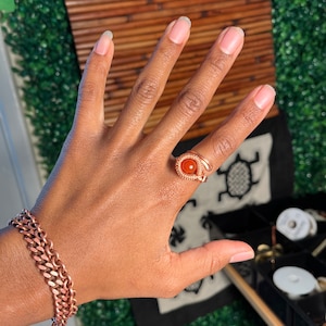 May include: A copper-colored ring with an oval, orange gemstone and a matching copper bracelet. The ring is on a hand with manicured nails. The bracelet has a chain link design.