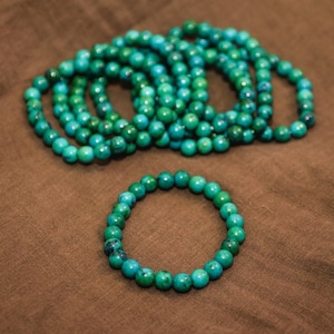 May include: A stack of turquoise green beaded bracelets. The bracelets are made of small, round beads and are all the same size. The bracelets are arranged in a pile, with the top bracelet being the most visible. The bracelets are on a brown fabric background.