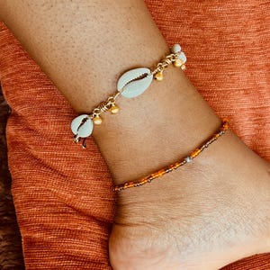 May include: Two anklets worn on the lower leg. The upper anklet features white cowrie shells interspersed with small gold-toned metal beads on a gold-toned chain. The lower anklet consists of a single strand of small orange and brown glass beads.