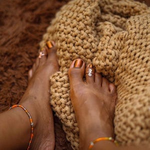May include: A silver-toned metal toe ring featuring a double spiral design worn on the second toe. The jewelry is paired with a thin, multi-colored beaded anklet and a thick, tan-colored chunky knit textile with a prominent woven texture.