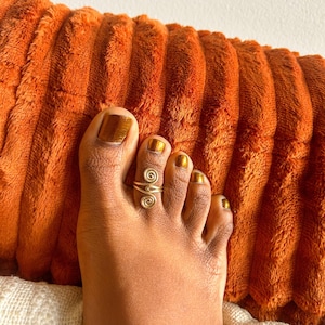 May include: A gold-toned metal toe ring featuring a double spiral design. The ring is worn on the second toe, showcasing a wire-wrapped aesthetic with two circular coils connected by a central band that wraps around the toe.