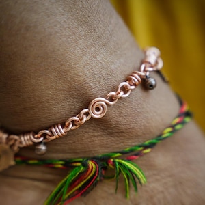 May include: A copper ankle bracelet featuring a spiral design and tiny bells. A colourful woven bracelet with red, yellow, green, and black threads is also visible. The jewellery is set against a blurred yellow background.