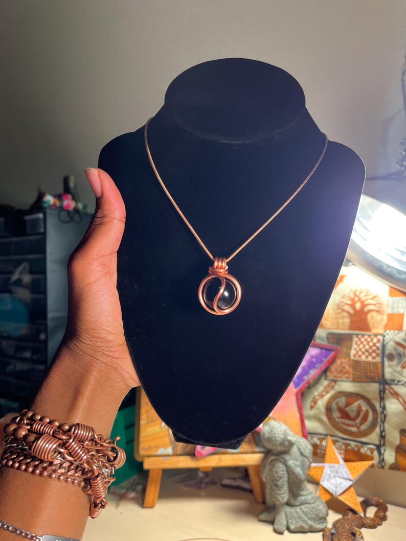May include: A copper wire wrapped necklace with a black gemstone pendant. The pendant is a sphere with a copper wire design.