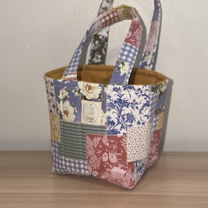 May include: A fabric tote bag featuring a patchwork design with various floral, gingham, and striped patterns in shades of blue, red, yellow, and white. The bag has two handles and a mustard yellow interior lining with a label reading The Cozy Stitch.