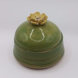 May include: A handmade ceramic jar with a lid, finished with a bright green glaze. The jar has a rounded form with horizontal ridges around the base. A yellow flower decorates the top of the lid, adding a decorative element. The jar is set against a plain white background.