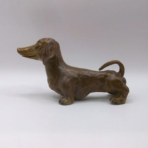 May include: A brown dachshund ornament. The dog is depicted standing, with its head raised and tail curled. The figurine has a smooth, matte finish and is set against a white backdrop. Dachshunds are a popular breed in the UK.