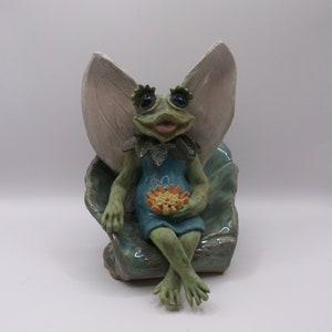 May include: A green frog fairy figurine with wings, sitting on a blue and green leaf-shaped chair, holding a yellow flower.