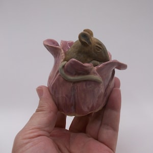 May include: A ceramic sculpture of a brown mouse nestled inside a pink flower. The flower has detailed petals and a green stem. The mouse is in a curled position, adding a whimsical touch to the piece. The sculpture is held in a hand.