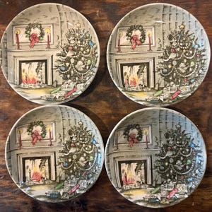 May include: A set of four round ceramic plates featuring a printed illustration of a cozy living room scene with a decorated Christmas tree, a fireplace with a lit fire, a festive wreath, and wrapped gifts stacked on the floor.