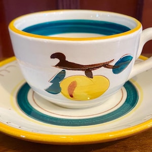 May include: White ceramic teacup and saucer set with a hand-painted design. The cup and saucer have a yellow rim and a teal and yellow painted design. The cup features a brown and yellow fruit design. The cup has a white handle.