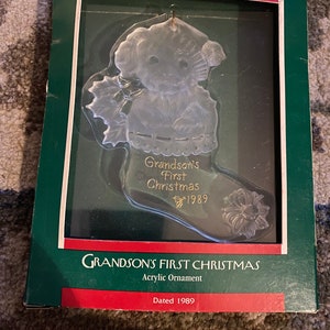 May include: Clear acrylic Christmas ornament in the shape of a stocking, featuring a teddy bear design. The ornament is labeled "Grandson's First Christmas 1989." The ornament is in a green and red box with the text "Grandson's First Christmas Acrylic Ornament Dated 1989."