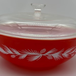 May include: A bright orange round glass casserole dish with a clear glass lid featuring a knob handle. The exterior of the bowl is decorated with a white horizontal pattern of pine needles and pinecones wrapping around the entire circumference.