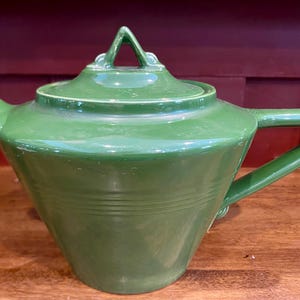 May include: A green ceramic teapot with a lid and handle. The teapot has a triangular handle on the lid and a curved handle on the side. The teapot is sitting on a wooden surface.