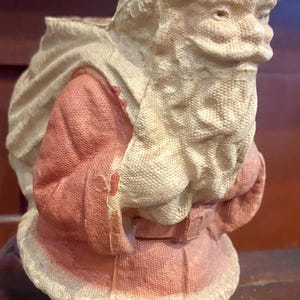 May include: A vintage Santa Claus figurine, featuring a rosy pink coat and hat with white trim. The figure carries a sack and has a detailed, textured appearance, evoking a classic holiday aesthetic.