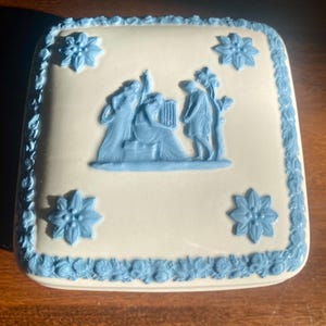 May include: Square, off-white ceramic trinket box with a blue bas-relief design. The lid features a classical scene with figures and floral accents. The box is trimmed with a blue floral border and blue flower accents.