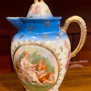 May include: Antique porcelain pitcher with a lid. The pitcher features a hand-painted scene of two people in a garden, with gold accents and a blue rim. The handle and lid finial are also gold. Pink floral details adorn the pitcher.