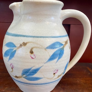 May include: Cream-colored ceramic pitcher with a curved handle and spout. The pitcher is decorated with blue leaves and pink flower buds on brown stems, with blue stripes near the top and bottom. A rustic, handmade aesthetic.