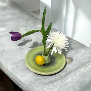 May include: A still life arrangement featuring a purple tulip, a white chrysanthemum, and a yellow lemon. The flowers are in a small vase set on a green ceramic plate. The plate has a decorative blue and green design.