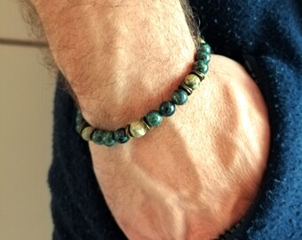 Puede incluir: Una pulsera de cuentas con cuentas de color verde azulado oscuro y crema. La pulsera está ensartada en un cordón flexible y acentuada con espaciadores de metal color bronce. La pulsera se lleva en la muñeca.