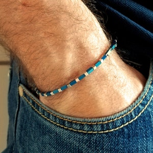 Peut inclure: Un bracelet bleu et argent avec des perles carrées. Le bracelet est au poignet d'une personne.