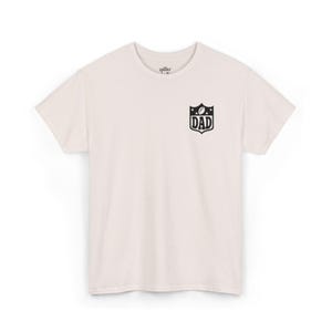 Può includere: T-shirt color crema con grafica nera sulla parte superiore sinistra del petto. La grafica è a forma di scudo con un pallone da football, stelle e la scritta "DAD". La maglietta ha maniche corte e girocollo.