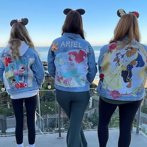 May include: Three denim jackets decorated with Disney characters. The jackets feature Ariel from The Little Mermaid, Belle from Beauty and the Beast, and Moana from Moana. Each jacket has a different design and color scheme.