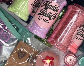 May include: A collection of small jars filled with colorful glitter, labeled "Maddie Belle Co." with different colors like green, purple, pink, and red. The jars are surrounded by various stickers with phrases like "Good Vibes" and "Today is a good day for a good day".