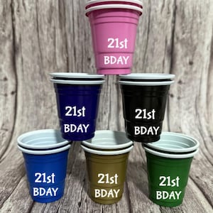 May include: A stack of six colorful plastic cups. The cups are pink, navy blue, black, blue, brown, and green. Each cup has a white rim and the text "21st BDAY" in white. The cups are arranged on a wooden surface.