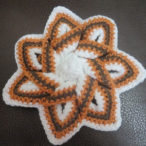Puede incluir: Un posavasos de ganchillo hecho a mano con forma de estrella. El posavasos presenta un centro blanco, bordeado por secciones marrones y naranjas, y delineado en blanco. El diseño es intrincado y detallado, mostrando la textura del hilo.