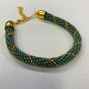 May include: A turquoise and gold beaded bracelet with a gold clasp. The bracelet is made of small, round beads that are woven together in a spiral pattern.