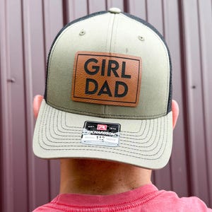 May include: A khaki and black trucker hat with the words "GIRL DAD" on a brown leather patch. The hat has a mesh back and a curved brim. The hat is a Richardson 112.