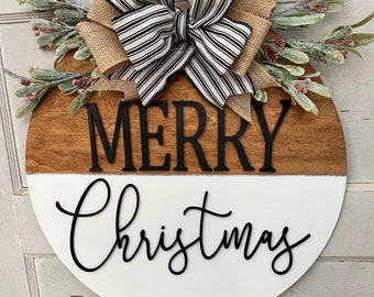 May include: A round wooden Christmas sign, split horizontally with a brown top and white bottom. The words "MERRY Christmas" are in black script. A burlap and striped bow and greenery with red berries adorn the top.