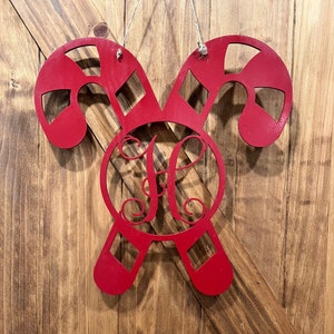 May include: Red wooden candy cane door hanger with a monogrammed circle in the center. The monogram is the letter "H" in a cursive font.