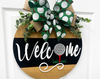 May include: Round wooden welcome sign with a golf theme. The sign features the word "Welcome" in white, a golf ball on a tee, and a green and white golf ball patterned bow. The sign is decorated with greenery.